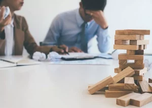 Deux collègues semblent en réunion dans un bureau, légèrement flous à l’arrière-plan. L’un d’eux tient sa tête entre ses mains, exprimant du stress ou de la fatigue, tandis que l’autre paraît parler ou expliquer quelque chose. Au premier plan, une tour en bois de type Jenga est partiellement effondrée, avec plusieurs blocs tombés sur la table. L’image symbolise la pression au travail, l’instabilité ou une situation professionnelle fragile.