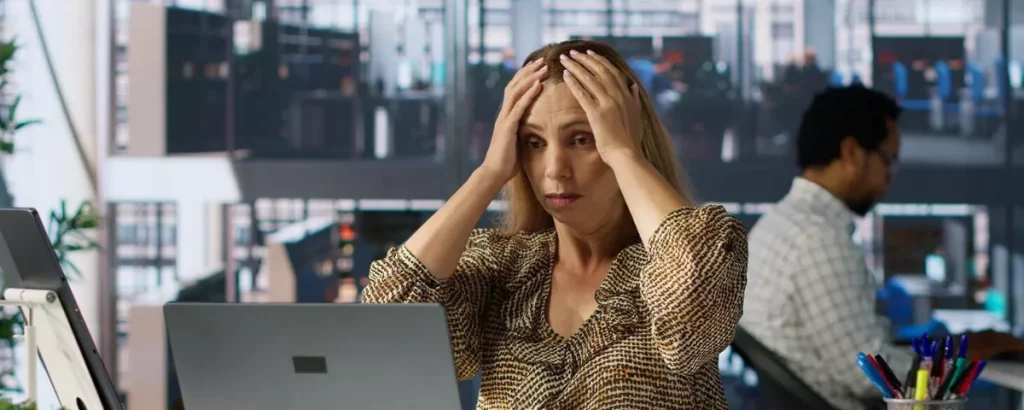 Une femme assise devant un ordinateur dans un bureau ouvert tient sa tête entre ses mains avec une expression inquiète et fatiguée. À l’arrière-plan, un collègue travaille à son poste. L’image évoque le stress professionnel, la surcharge de travail ou l’anxiété au bureau.
